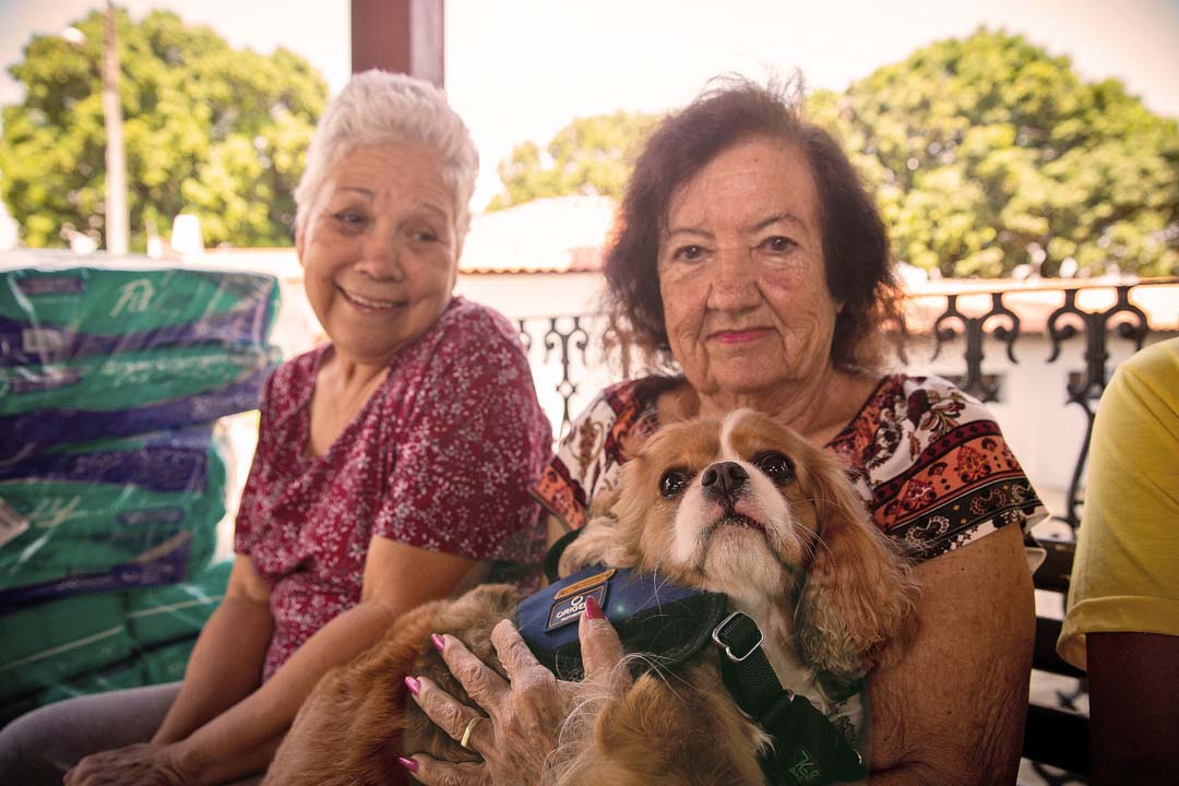 Na imagem um cão no colo de uma idosa durante a visita em um lar de idosos, enquanto outra idosa ao seu lado está sorrindo olhando para o cão.
