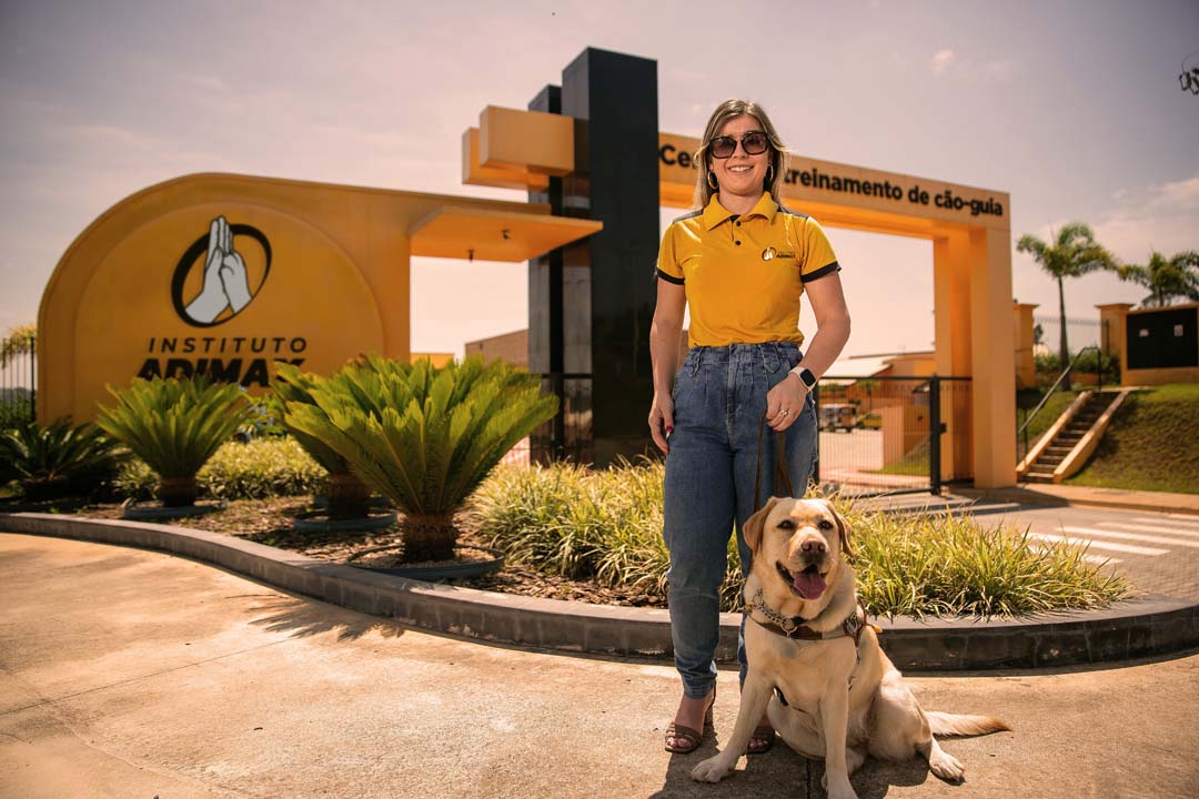 Imagem de uma mulher com seu cão-guia em frente ao Centro de Treinamento do Instituto Adimax