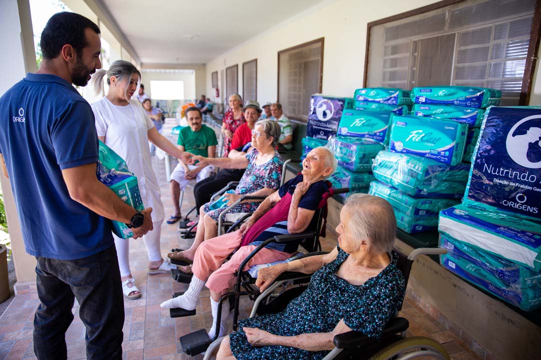 Na imagem, um homem carregando um pacote com fraldas geriátricas doado pelo Instituto Adimax e vários idosos sentados conversando com o mesmo.