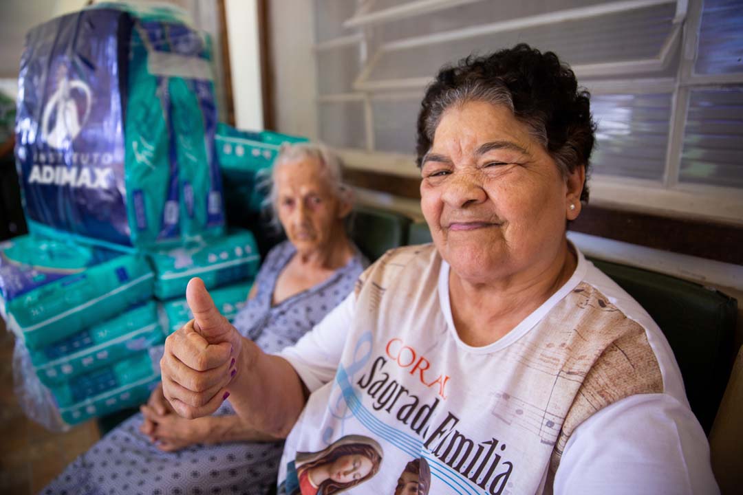 Na imagem, duas idosas, uma acenando com positivo e ao fundo várias fraldas geriátricas doadas pelo Instituto Adimax.