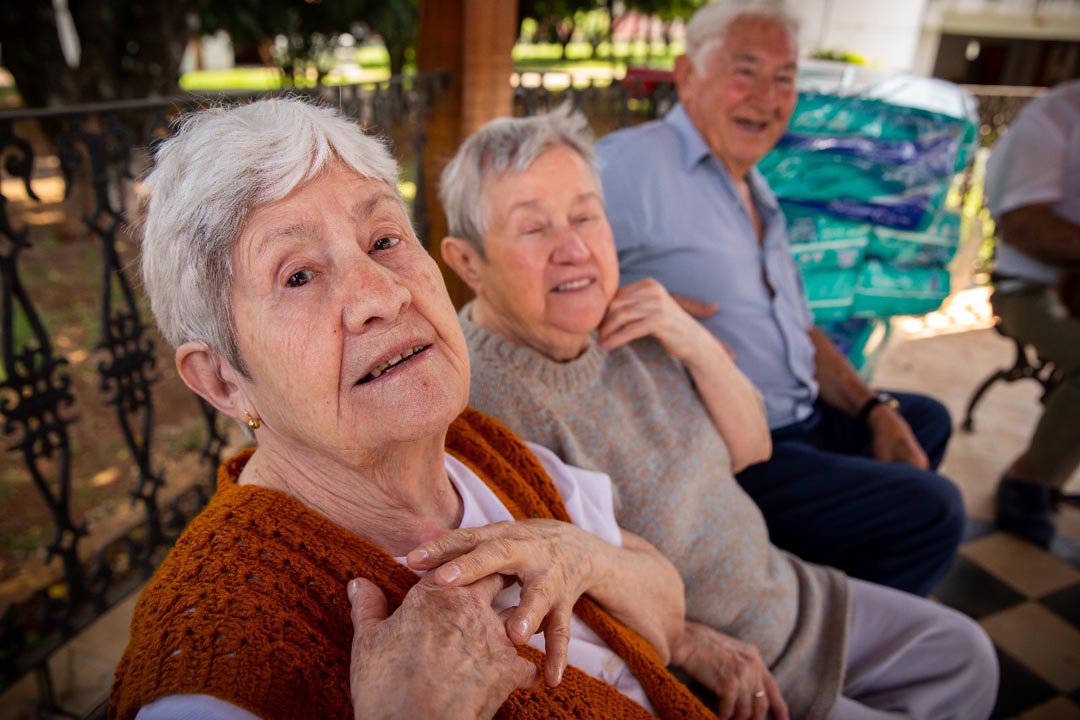Na imagem 3 idosos, um deles olha para a câmera com as mãos em seu peito 