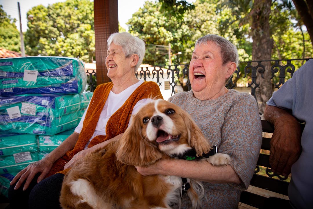 Na imagem o cão da Pet Terapia está no colo de uma senhora idosa enquanto outra está sentada ao lado, ambas estão sorrindo