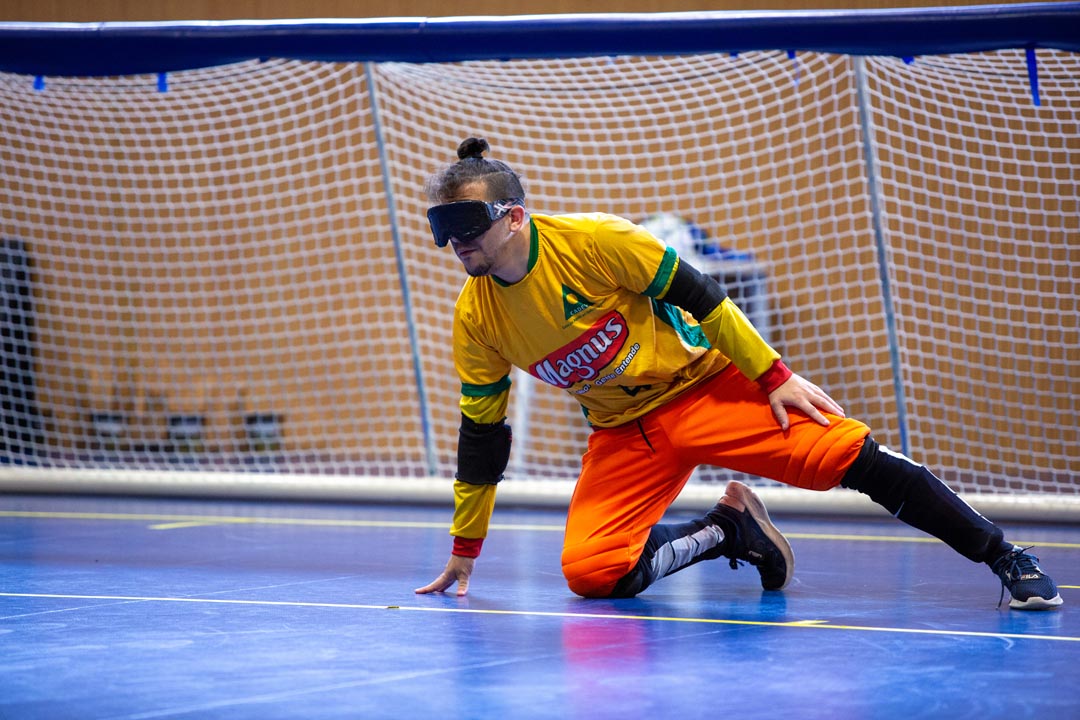 Na imagem atleta do Goalball apoiado com o joelho e uma mão, durante uma partida, vestindo uniforme da Magnus e venda nos olhos