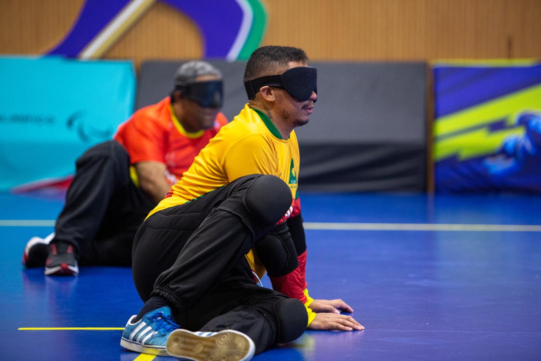 Na imagem dois atletas do Goalball sentados no chão enquanto estão em uma partida, ambos vestem uniforme Magnus e utilizam vendas nos olhos