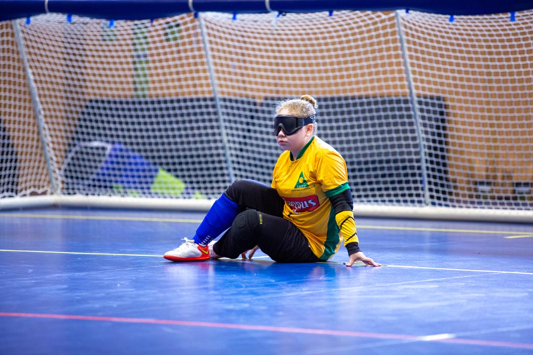 Na imagem atleta do Goalball sentada no chão e se apoiando em uma mão, durante a partida, veste uniforme Magnus e venda nos olhos
