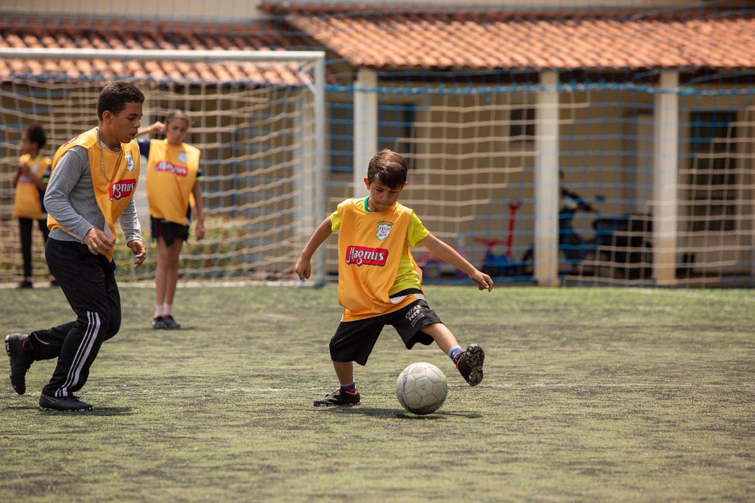 Na imagem menino da organização assistencial joga a bola em um campo de futebol, e veste coleta Magnus. Mais 3 crianças participam do jogo.