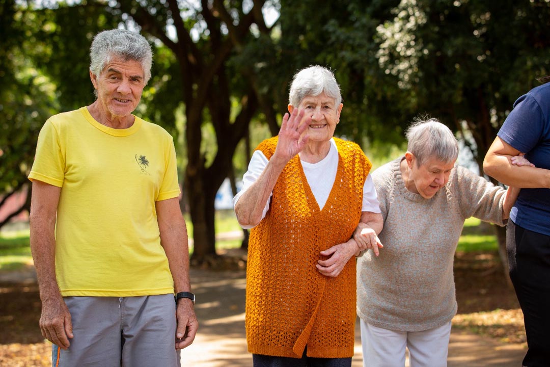 Na imagem 1 homem idoso e duas mulheres idosas segurando o braço uma da outra, uma delas olha e da tchau pra câmera