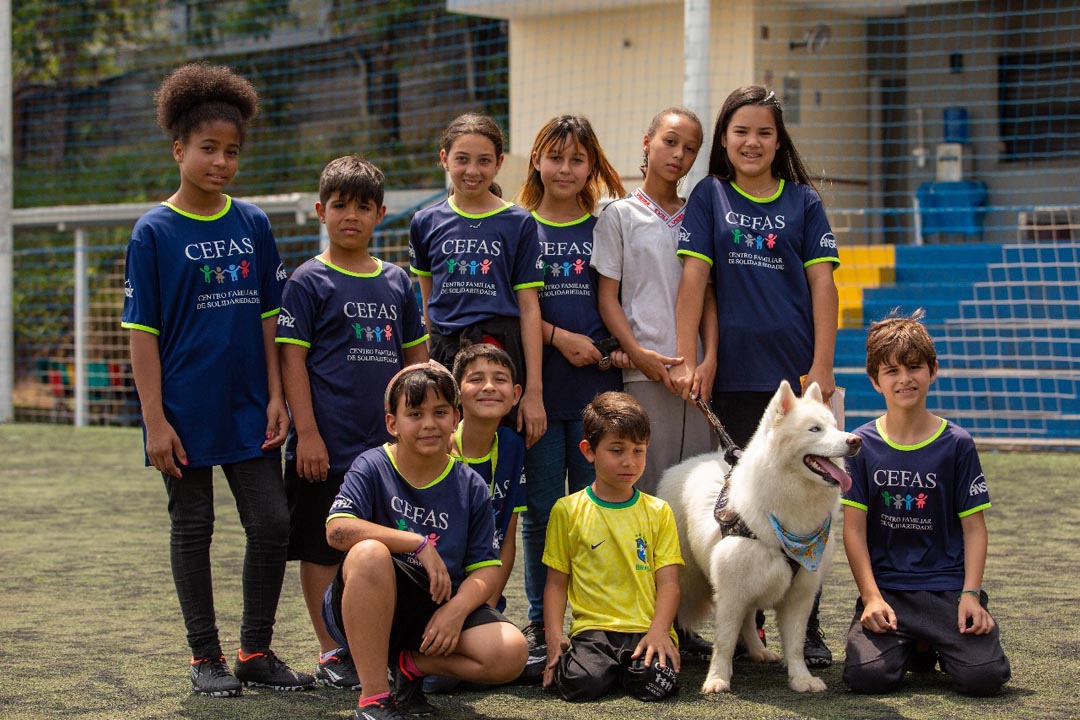Na imagem 10 criança da organização assistencial CEFAS olham para foto em um campo de futebol, acompanhado de um cachorro raça pastor alemão, cor branco.