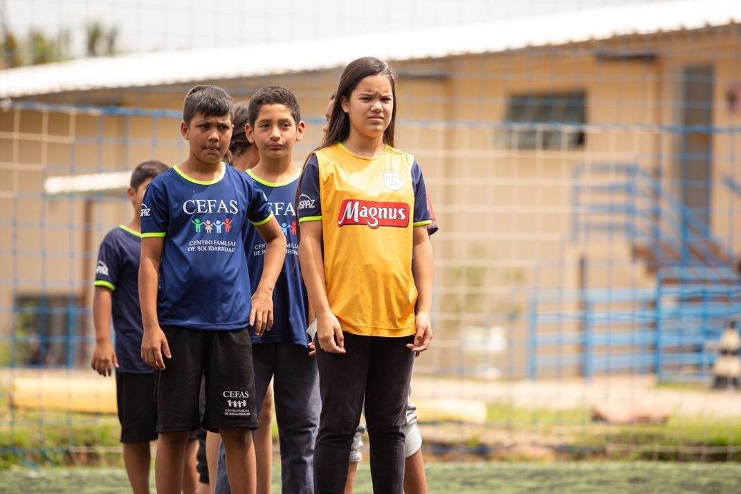 Na imagem uma fila de 6 alunos com uma menina na frente vestindo colete Magnus, em um campo de futebol, todos da organização assistencial CEFAS